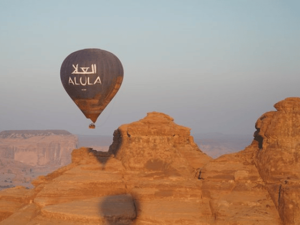 Hot air balloon with 'AlUla' logo floating over ancient rock formations in AlUla, Saudi Arabia at sunrise.