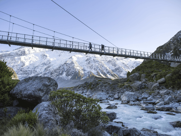 Hikers crossing a suspension bridge overlooking a glacial river and snow-capped mountains in Aoraki-Mount Cook National Park, New Zealand