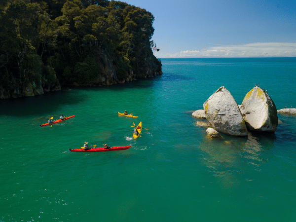 Kayakers paddling around Split Apple Rock in Abel Tasman National Park, New Zealand. The water is a vibrant turquoise color and the surrounding landscape is lush green