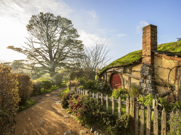 Hobbiton, Waikato_ Charming Hobbiton house with a red door, surrounded by a picturesque garden and picket fence, with a large tree in the background, located in Waikato